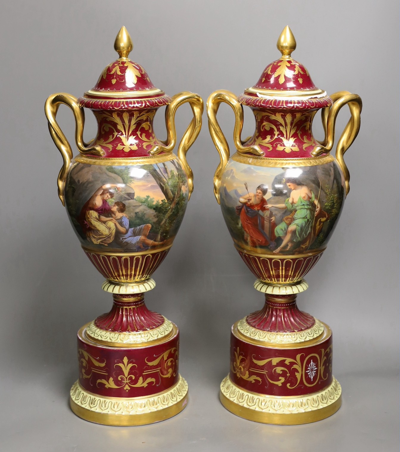 A pair of ornate Vienna style two handled urns bearing covers, with painted love scene and red ground and gilt decoration, 46.5cm high